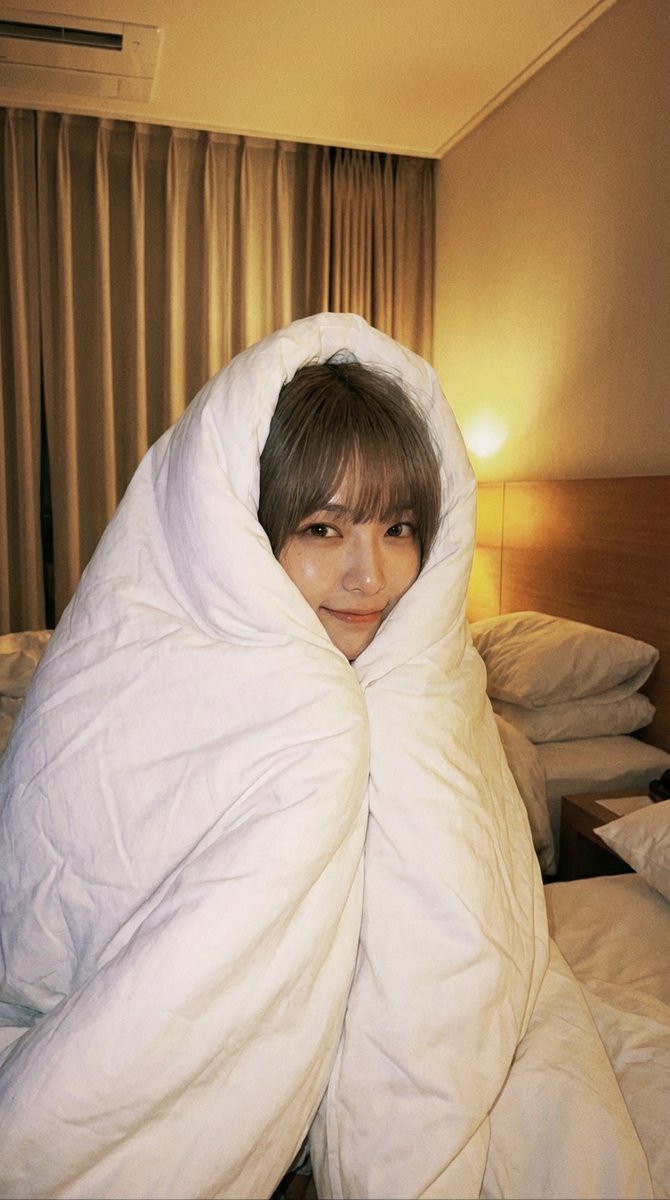 A young Asian woman wrapped in a white duvet, face peeking out, cozy candid selfie style