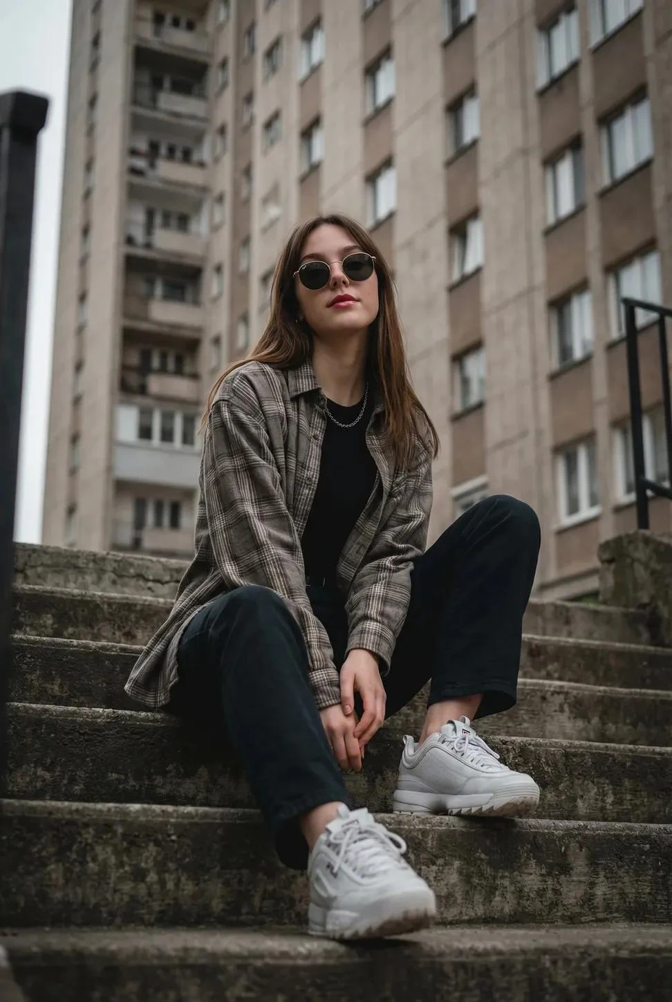 A young woman sitting casually on outdoor concrete stairs, posing with a cool, confident demeanor.