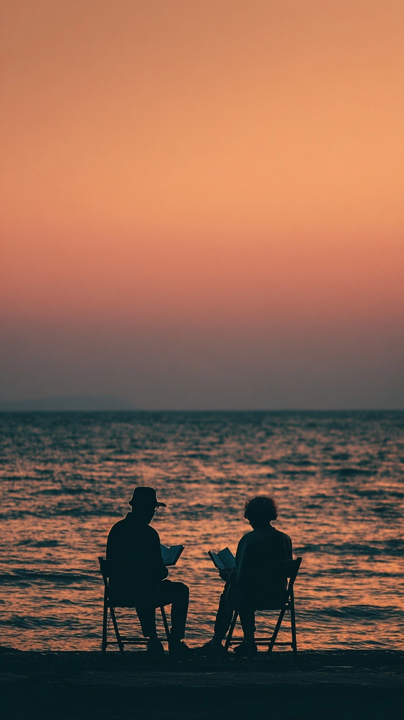 A man and woman sitting on chairs by the sea, reading books, with their backs to us, at dusk