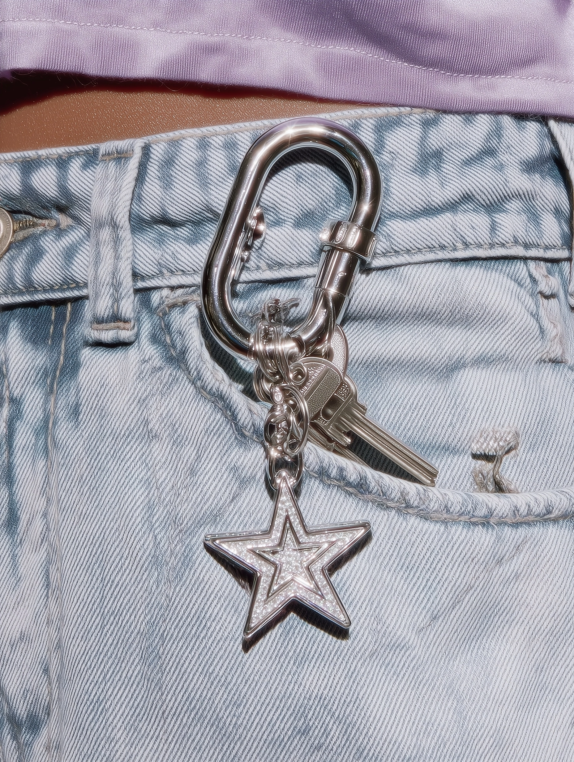 An editorial close-up shot of a star-shaped metal carabiner clipped onto a light blue denim belt loo