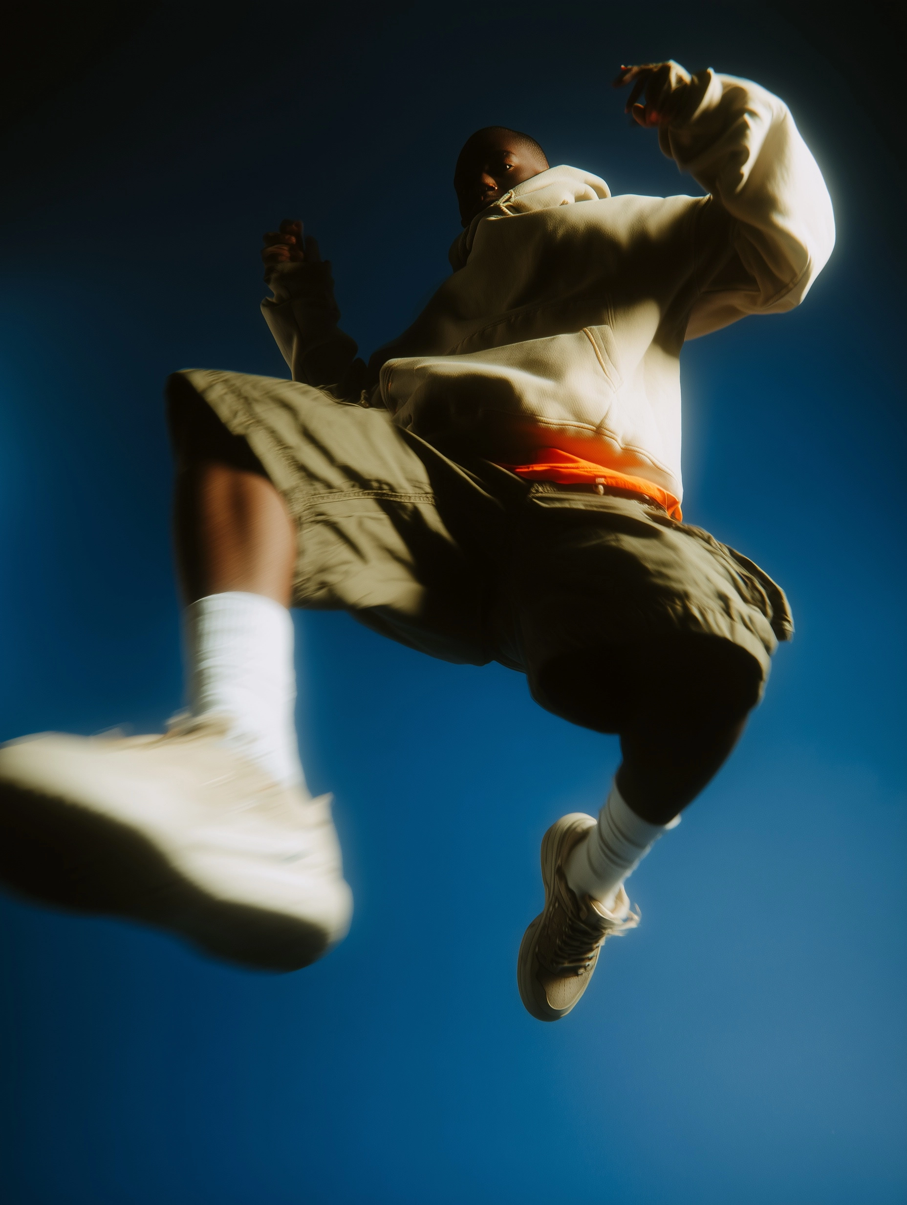 Capture a dynamic mid-air jump of a dark skinned person wearing a khaki hoodie and shorts with white