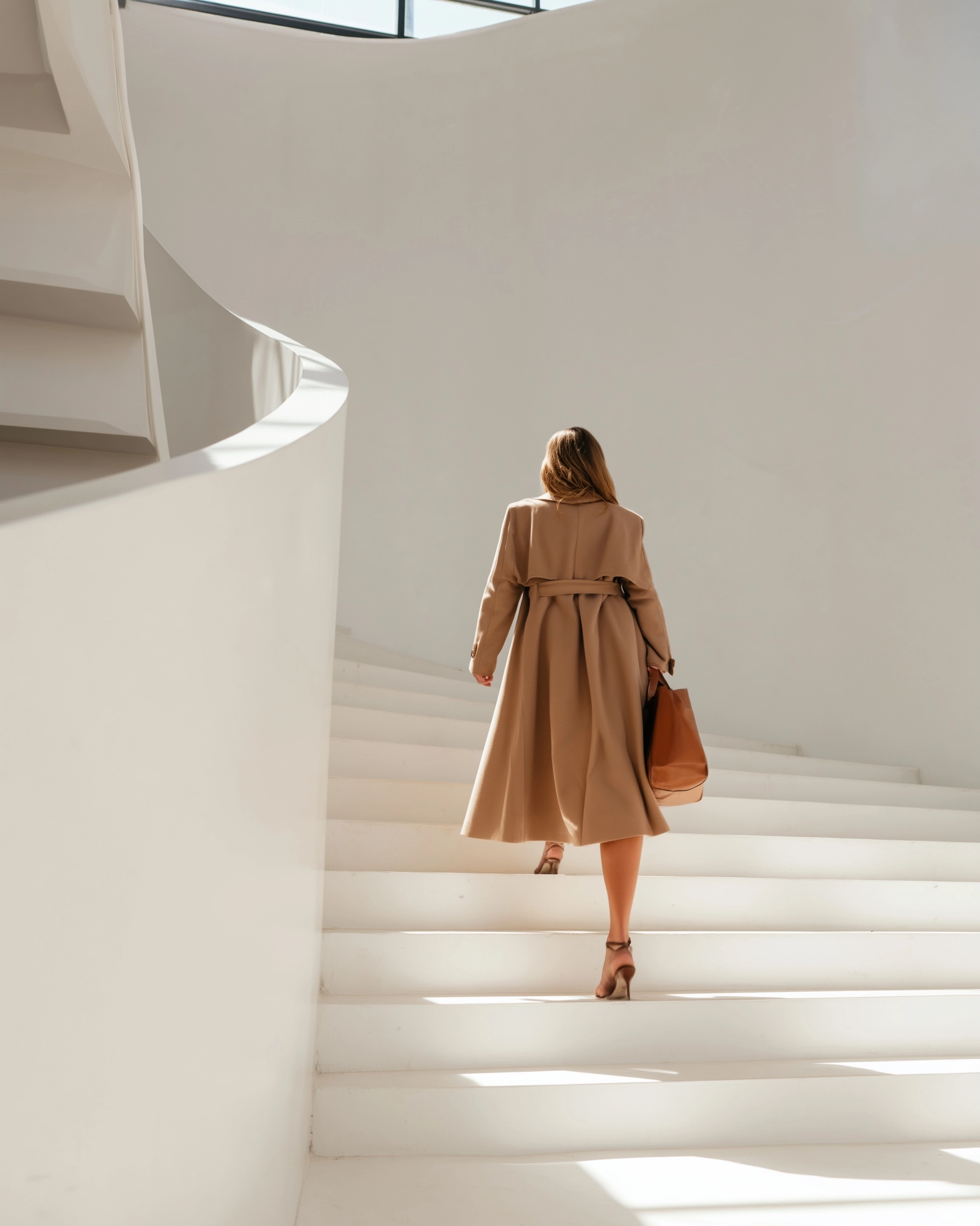Editorial cinematic wide shot of a young woman from behind walking up a sweeping minimal white stair