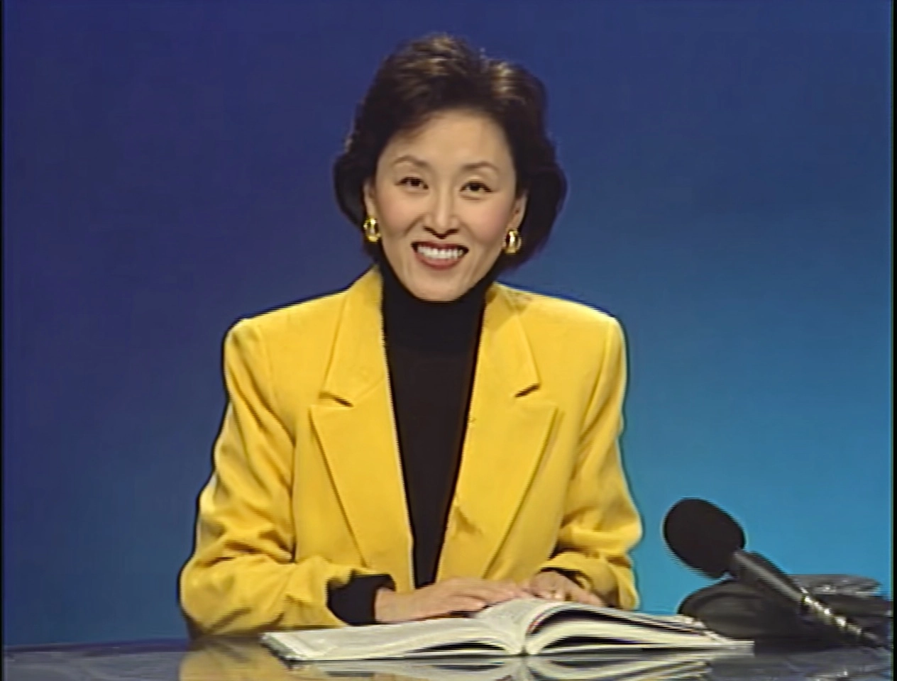 A chinesefemale news anchor, sitting at a desk in front of a microphone and speaking into it with bo