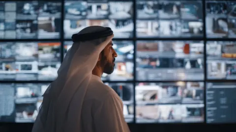 Traditional Emirati attire stands in the center, seen from behind. In front of him is a large wall o