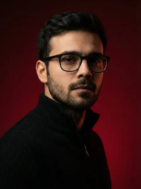 A man with dark hair and glasses wearing a black knit zip-up sweater against a deep red background.