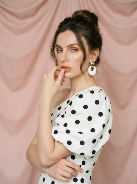 A woman in a white dress with black polka dots and a single puffed sleeve, wearing large white and gold earrings, with her hand resting near her lips against a pink draped fabric.