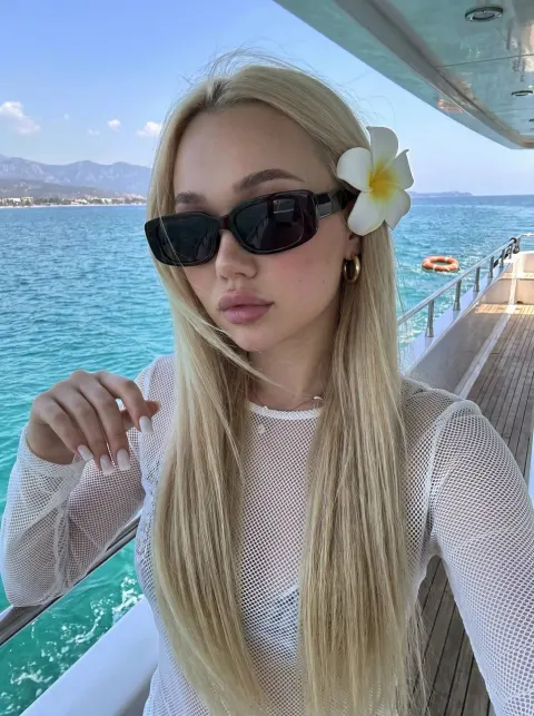 A blonde woman wearing black sunglasses and a white mesh top with a flower in her hair on a boat.