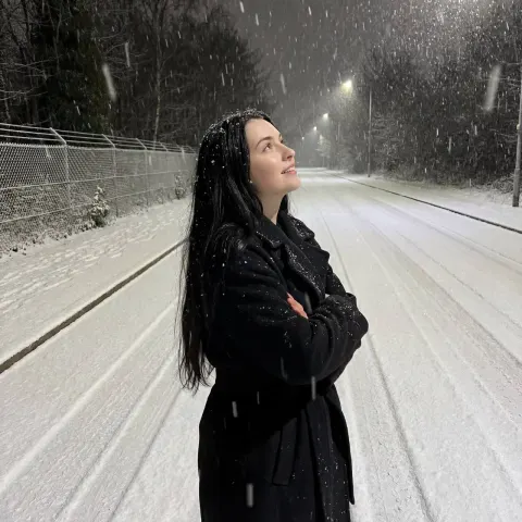 A woman with long dark hair stands in a snowy road at night wearing a black coat.
