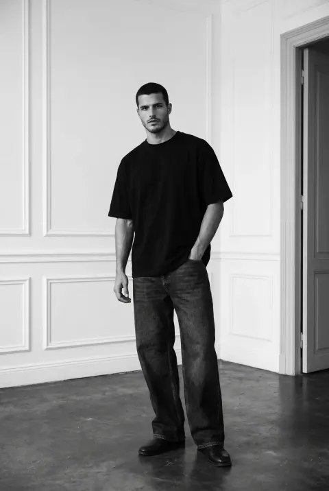 A black and white full-body portrait of a man in a minimalist room. He wears a plain black t-shirt, dark wide-leg jeans, and dark shoes, standing with one hand in his pocket.