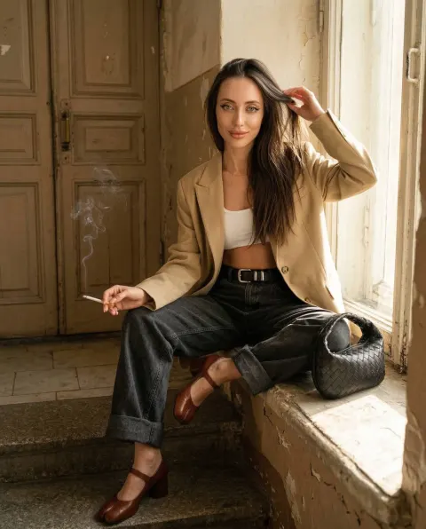 A woman with long dark hair sits on a windowsill, holding a lit cigarette, in a sunlit hallway with a weathered door behind her.