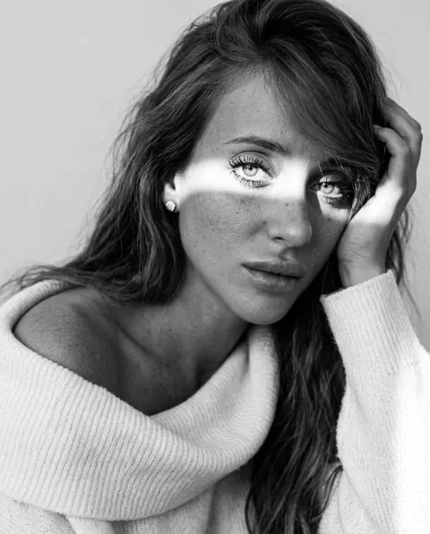 A black and white portrait of a person lying down, looking into the camera with a hand supporting the temple. A horizontal strip of bright light illuminates the eyes and bridge of.