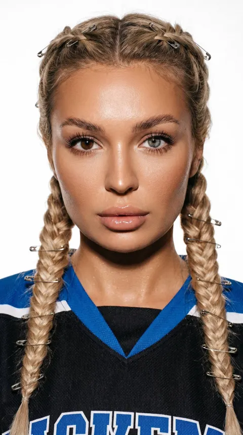 Close-up portrait of a person with two braids adorned with silver safety pins, wearing a black and blue jersey, against a white background. The subject has heterochromia, with one.