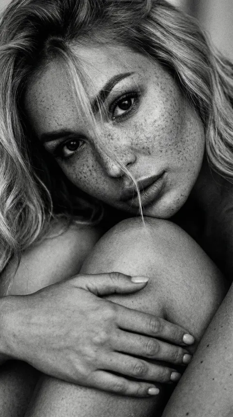 A black and white extreme close-up portrait of a person with freckles, looking directly at the camera. They are hugging their bent knee, with loose hair falling across their face.