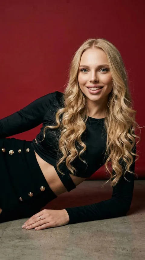 A woman with long, wavy blonde hair reclines on a concrete floor against a solid red background, wearing a black long-sleeved top with a stomach cutout and a black skirt with gold.