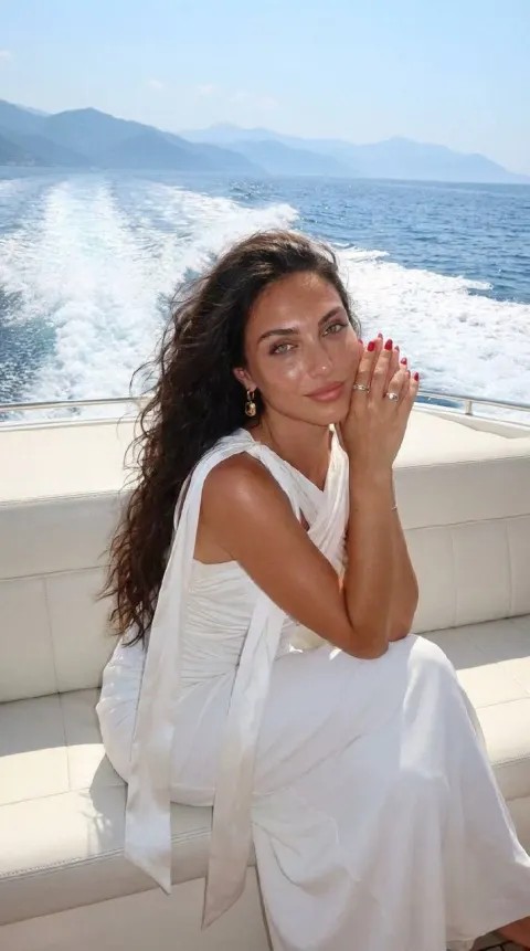 A woman with long dark hair wearing a white draped gown sits on a boat deck overlooking blue water.