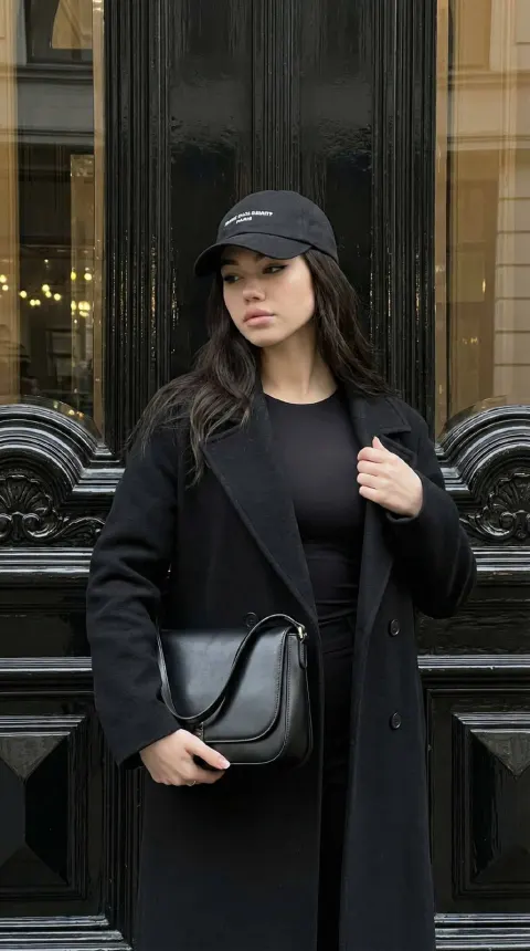 A person in a black coat and cap stands against a large, ornate dark door.