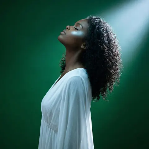 A person with curly hair in a white dress stands against a deep green background, head tilted back.
