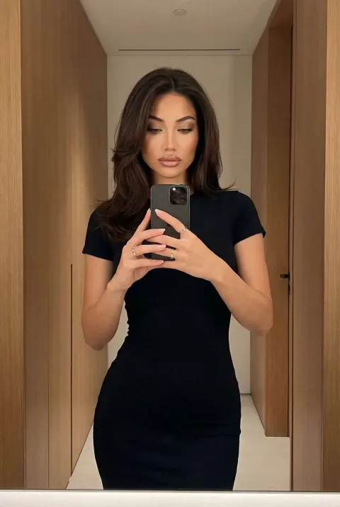 A woman in a fitted black short-sleeve dress takes a mirror selfie inside a modern room with wooden paneling.