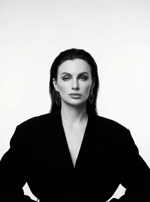 A woman in a black jacket with slicked-back hair poses against a light gray background.