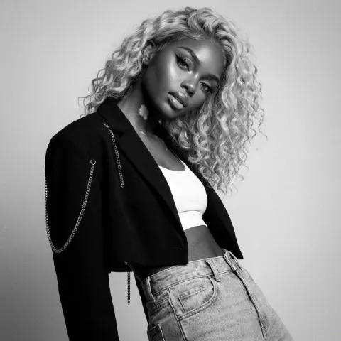 A person with long curly hair wearing a cropped black jacket adorned with silver chains over a white top and light blue jeans.