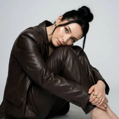 A woman in a dark brown leather blazer and matching trousers sits on the floor with her knees pulled to her chest.