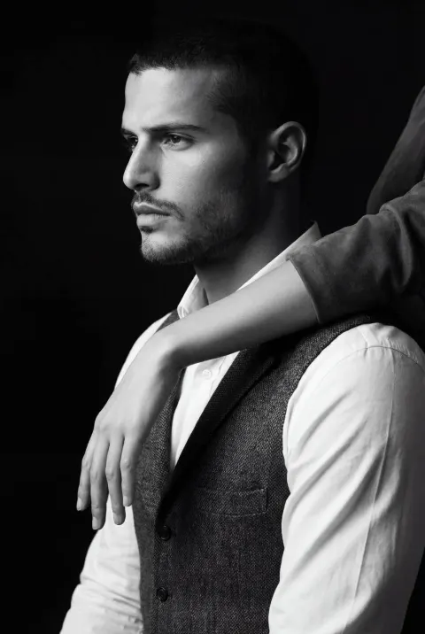 A black and white medium shot shows a man in profile wearing a vest and shirt with a woman's arm resting across his shoulder.