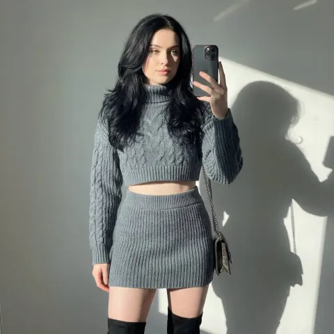 A person with long dark hair wearing a grey cropped cable knit sweater and matching ribbed mini skirt stands against a light wall.
