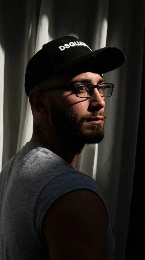 A man wearing a black baseball cap and glasses stands against a backdrop of draped curtains.