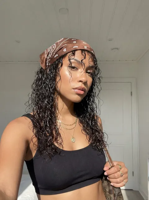 A person with curly dark hair wears a brown patterned bandana and a black crop top while holding a small bag.