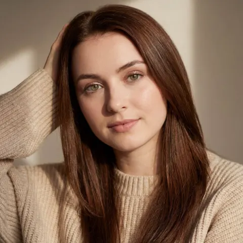 A woman with long brown hair wearing a light beige ribbed sweater poses against a neutral background.