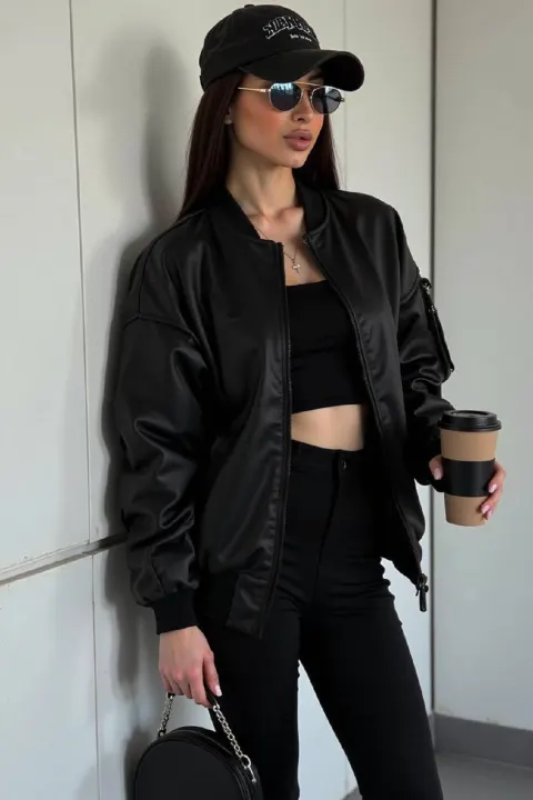 A woman in a black bomber jacket and crop top stands against a white wall holding a coffee cup.