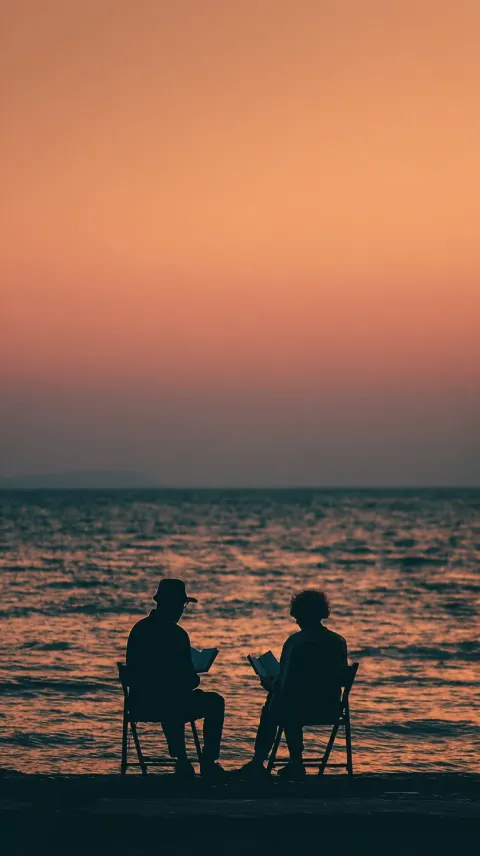 A man and woman sitting on chairs by the sea, reading books, with their backs to us, at dusk