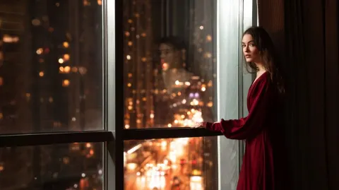A young woman stands by the window of her high-rise apartment