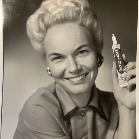 A photo of a 1950s housewife, smiling and holding up a tube of "lady glue"