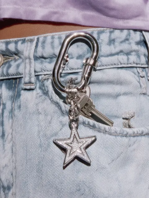 An editorial close-up shot of a star-shaped metal carabiner clipped onto a light blue denim belt loo