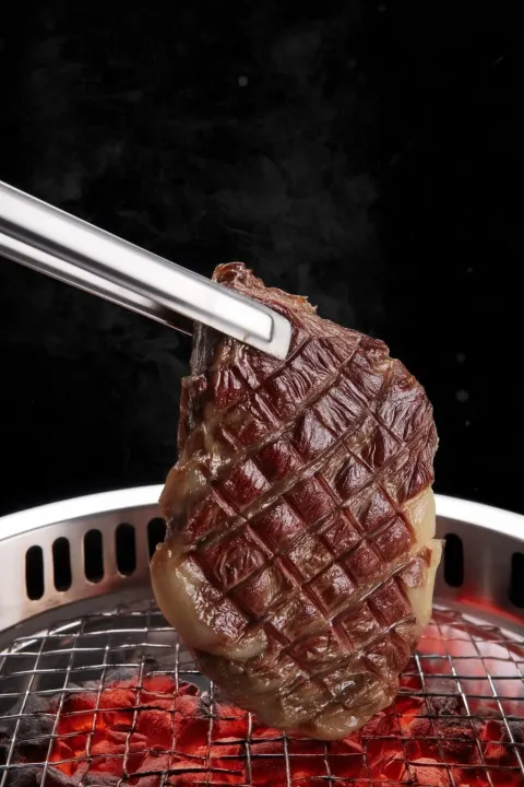 Extreme close-up of stainless steel long tongs gripping a thick premium wagyu beef steak with deep c