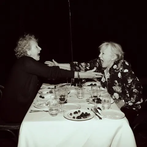 Two elderly women in their 80s screaming and lunging at each other across a formal fine-dining table