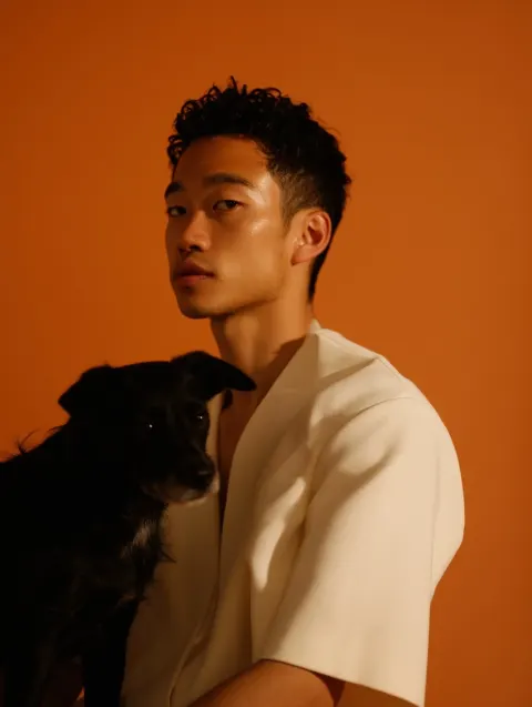 Three-quarter portrait of a young white Asian man with a black dog, warm orange-white background
