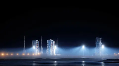 Wide night shot of rocket launch complex, engine plume lighting entire launch pad