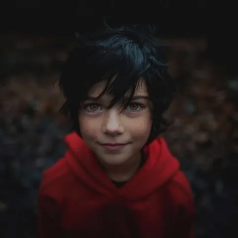 A cryptid nine year old boy in a red hoodie, amber eyes, black messy hair, horror child, smile